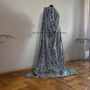 Può includere: Un mantello lungo fino a terra realizzato in tessuto argentato riflettente con un motivo olografico a vetro incrinato che brilla di luci multicolori. Il capo è drappeggiato su un manichino e riporta il testo https://www.etsy.com/shop/CoutureDivision.