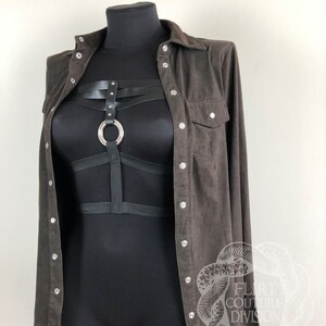 以下が含まれることがあります： 茶色のスエードシャツに黒いハーネスと銀色の金具が付いている。シャツはボタンダウンの前立てと長袖。ハーネスは中央にリングがあり、複数のストラップが付いている。
