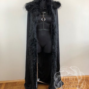 以下が含まれることがあります： 黒いフェイクファーのフード付きケープで、長く流れるようなデザイン。ケープは黒い生地で裏地が付けられており、ネックラインにシルバーの金属製のリングのディテールがあります。