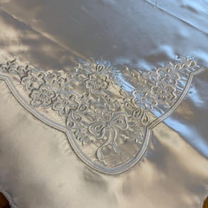May include: A white satin tablecloth with a scalloped edge and a detailed floral embroidery in light blue. The embroidery features flowers and a bow design, adding a touch of elegance to the fabric.