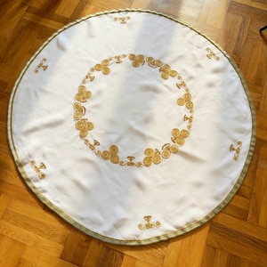 Può includere: Una tovaglia rotonda bianca con ricami dorati a spirale e floreali. La tovaglia ha un bordo dorato e verde. La tovaglia è su un pavimento di legno.