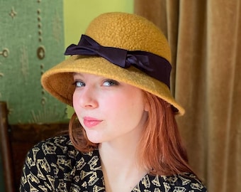May include: A mustard yellow felt cloche hat with a wide brim and a black satin ribbon tied in a bow at the front.