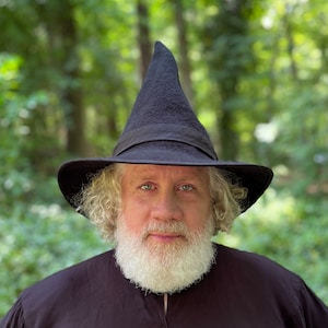 Op de afbeelding: Een man met een lange witte baard die een zwarte, puntige hoed met een brede rand draagt. Hij staat voor een wazige groene achtergrond.