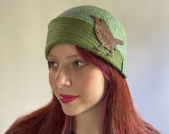 May include: A woman wearing a green felt cloche hat with a brown felt bird applique. The hat has a wide brim and a soft, slouchy shape.
