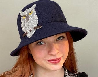 May include: A navy blue felt bucket hat with a gray owl applique. The owl has yellow eyes and brown accents.