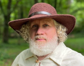 May include: A man with a long white beard and curly hair is wearing a brown felt hat with a wide brim and a leather band. He is wearing a light beige shirt with wooden buttons.