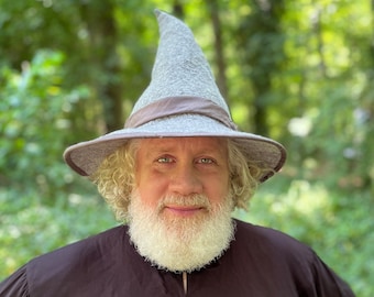 May include: A man with a long white beard is wearing a gray pointed hat with a brown band. He is looking directly at the camera and has a friendly expression.