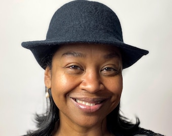 May include: A black wool cloche hat with a rounded crown and a small brim. The hat is worn by a person with dark hair and a smiling expression. The background is a plain white.