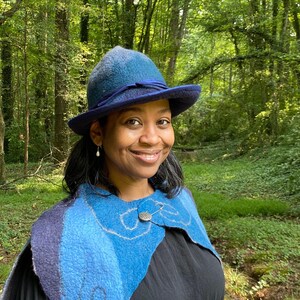 May include: A woman wearing a blue hat with a blue and white patterned scarf. She is standing in a woodland setting.