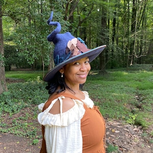 May include: A woman wearing a blue, brown, and green felt witch hat with a pointed crown and a wide brim. The hat is decorated with felt leaves and flowers. She is wearing a brown dress with a white lace overlay.