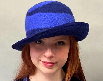 Puede incluir: Un sombrero azul con ala, con rayas horizontales en diferentes tonos de azul. El sombrero tiene una copa redondeada y un ala ligeramente inclinada. El sombrero está hecho de un material suave y texturizado.