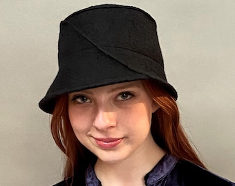May include: A black felt bucket hat with a unique angled seam design. The hat is worn by a person with red hair.