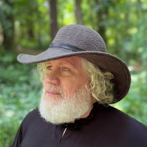 May include: A man with a long white beard wearing a brown felt hat with a black leather band. The hat has a wide brim and is slightly tilted to the side.