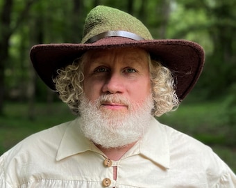 May include: A man with a white beard is wearing a brown and green felt hat with a brown leather band. He is wearing a white shirt with brown buttons.