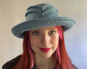 Puede incluir: Un sombrero de fieltro azul claro con un ala ancha y un diseño único en capas. El sombrero tiene un acabado suave y texturizado y un ala ligeramente curvada.