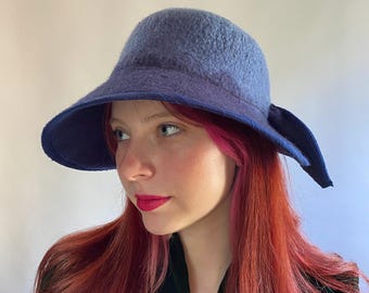 Puede incluir: Un sombrero de fieltro azul con un ala ancha y un lazo en un lado. El sombrero está hecho de un material suave y texturizado.