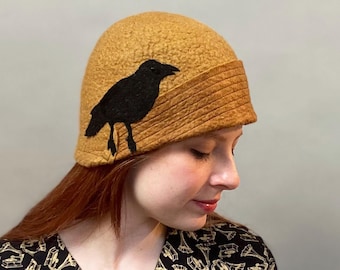 May include: A mustard yellow felt cloche hat with a black felt crow applique. The hat has a wide brim and a fitted crown.