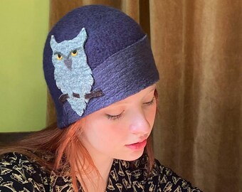 May include: A dark blue felt cloche hat with an owl applique. The owl is gray with yellow eyes and is perched on a branch.