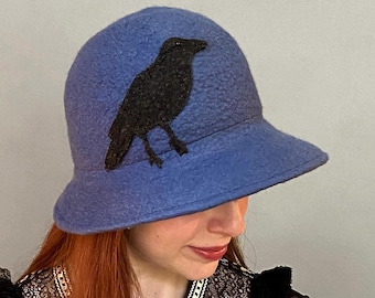 May include: A blue felt cloche hat with a black felt crow applique. The hat has a wide brim and a rounded crown.