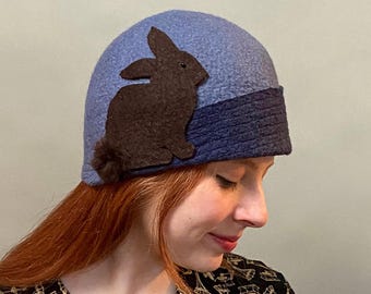 May include: A blue and navy blue felt cloche hat with a brown felt rabbit applique. The hat is worn by a person with red hair.