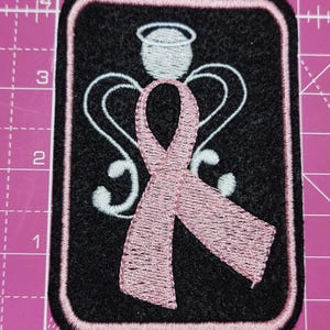 May include: Embroidered patch featuring a pink ribbon, angel wings, and a halo on a black background. The patch has a pink border. The design is likely related to breast cancer awareness or support for a cause.
