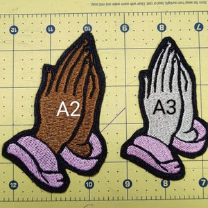 May include: Embroidered patches featuring praying hands in various color combinations. The patches include brown, tan, and white hands with pink and black accents. Each patch is labeled with a letter and number combination.