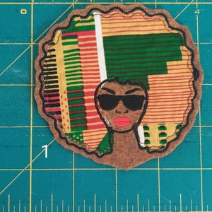 May include: A brown felt patch with a colorful patterned design. The design features a woman with black sunglasses and red lips. The pattern is made up of yellow, green, red, and black stripes.