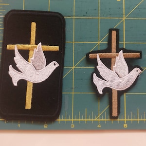 May include: Two embroidered patches featuring a white dove flying over a gold cross. The larger patch is on a black background. The smaller patch is on a brown background.