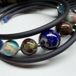 May include: A multi-strand wrap bracelet featuring a smooth black rubber cord accented with polished spherical beads in shades of deep blue, turquoise, brown, and green, separated by small silver-toned metal spacers along the flexible band.