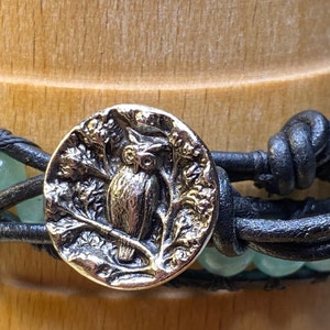 May include: Wrap bracelet with a silver owl button clasp. The bracelet features light green beads and black leather straps. The owl button is detailed with an owl perched on a branch, surrounded by foliage. The bracelet is wrapped around a wooden dowel.
