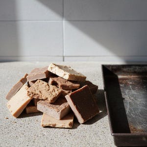 May include: A pile of square-shaped, multi-colored soap bars in various shades of brown, tan, and beige. The bars have different textures, ranging from smooth to porous and crumbly. A rectangular, dark metal tray sits to the right of the soap pile.