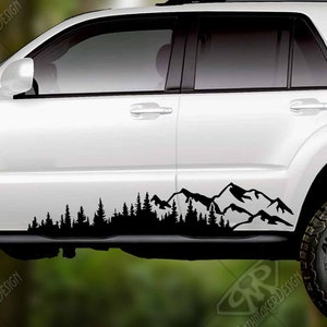 May include: A white SUV with black mountain and forest decals along the side. The tires are black with silver rims. The background is a blurred forest scene.
