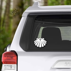 May include: White vinyl sticker of a scallop shell with a cross inside. The sticker is on the back window of a white car.