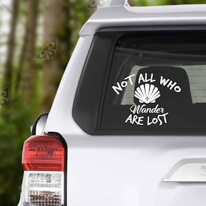 May include: White vinyl decal with the text "Not all who wander are lost" and a seashell with a cross inside. The decal is on the back window of a silver car.