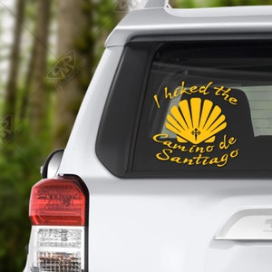 May include: A white car with a yellow sticker on the back window. The sticker says "I hiked the Camino de Santiago" and features a yellow scallop shell with a cross.