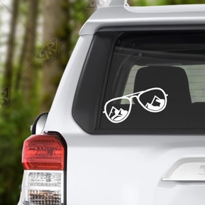 May include: White vinyl decal of a pair of sunglasses with mountains reflected in the lenses. The decal is applied to the rear window of a silver SUV.