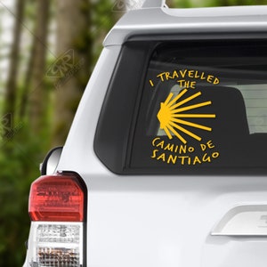 May include: Yellow vinyl decal on a car window. The decal reads "I TRAVELLED THE CAMINO DE SANTIAGO" with a stylized sunburst design. The decal is applied to the rear window of a white SUV.