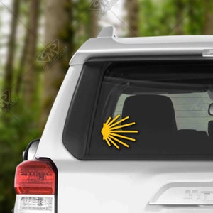 May include: A yellow scallop shell sticker on the back window of a white car. The sticker is a symbol of the Camino de Santiago pilgrimage route.