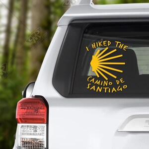 May include: A white car with a yellow sticker on the back window. The sticker says "I hiked the Camino de Santiago" and features a yellow scallop shell.