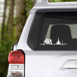 May include: White vinyl decal of a heartbeat with a seashell, cross, and church building. The decal is on the rear window of a silver car.
