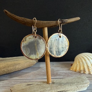 May include: A pair of circular earrings with a gold-coloured trim and a pearlescent, grey and white centre. The earrings are displayed on a wooden stand against a dark background. They have a hook closure.