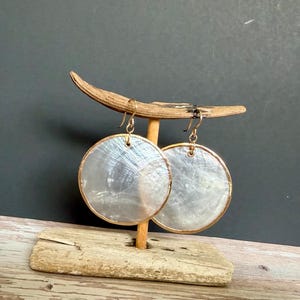 May include: A pair of circular earrings featuring iridescent white shell discs encased in thin gold-toned metal rims. The earrings hang from gold-toned metal hooks, displaying a natural pearlescent sheen and a smooth, polished surface texture.