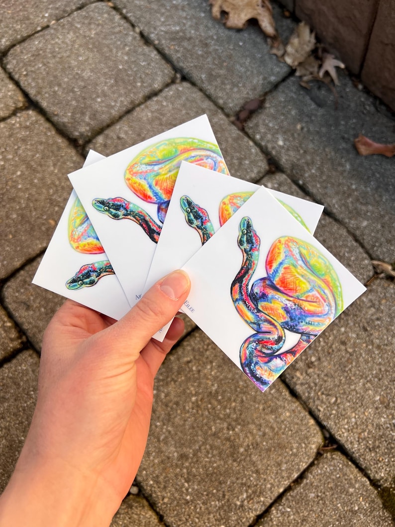 Op de afbeelding: Een hand houdt een stapel vierkante witte kaarten vast met kleurrijke slangenillustraties. De slangen zijn afgebeeld met levendige regenboogtinten, waaronder blauw, groen, geel en rood. Het kunstwerk is gedetailleerd en toont de schubben en patronen van de slang.