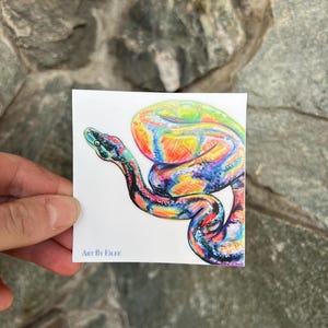 Op de afbeelding: Een kleurrijke sticker met een gestileerde slangenillustratie. De slang is afgebeeld met levendige tinten blauw, groen, geel en rood. De tekst "ART BY EILEE" staat onderaan de sticker.