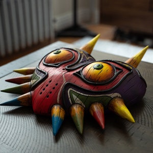 May include: A colorful Majora's Mask replica with sharp, pointed edges. The mask is painted in shades of red, yellow, green, blue, and purple. The mask has two large, yellow eyes with black pupils and green irises.