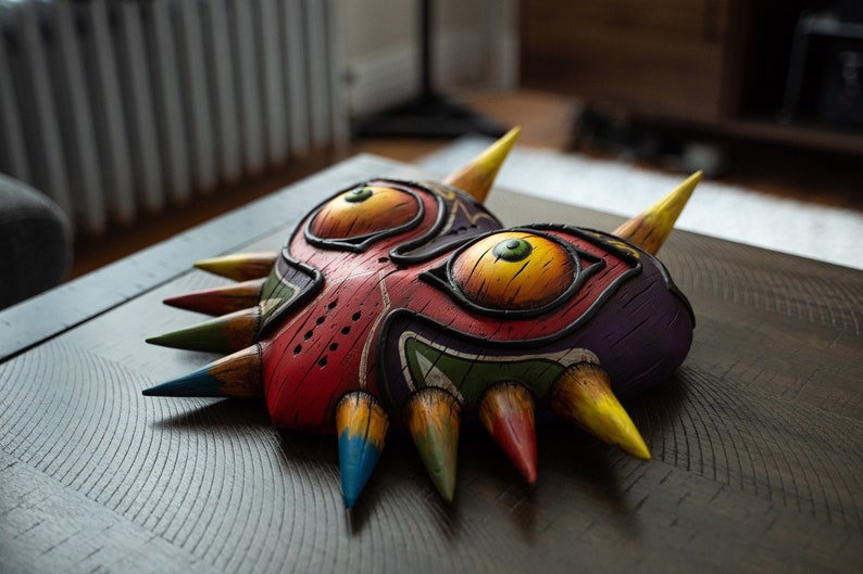May include: A colorful Majora's Mask replica with sharp, pointed edges. The mask is painted in shades of red, yellow, green, blue, and purple. The mask has two large, yellow eyes with black pupils and green irises.