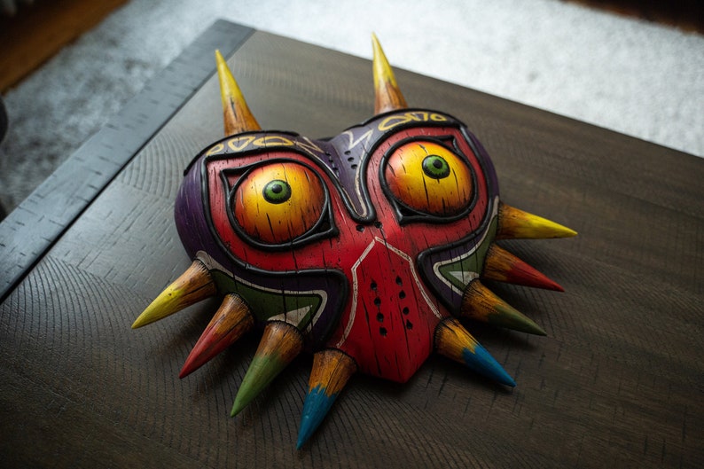 May include: A red, yellow, green, and purple wooden mask with a heart shape and sharp points. The mask has two large eyes with yellow pupils and green irises.