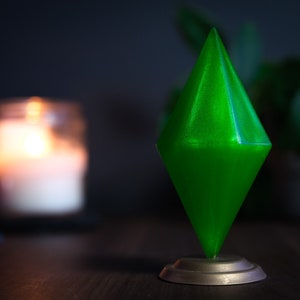 May include: A green diamond-shaped figurine with a shiny, iridescent finish. The figurine is on a silver base and is sitting on a wooden surface.