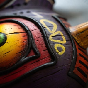 May include: Close-up of a wooden mask with a yellow, orange, and red eye. The mask is painted with black lines and has a purple background. The mask has a yellow and brown beak.