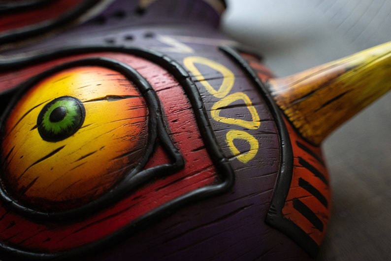 May include: Close-up of a wooden mask with a yellow, orange, and red eye. The mask is painted with black lines and has a purple background. The mask has a yellow and brown beak.